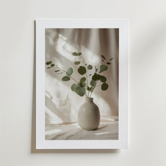 Eucalyptus Vase Poster från Bozetto föreställer en enkel keramikvas med gröna eukalyptusgrenar på en mjukt upplyst, neutral bakgrund, tryckt på FSC-certifierat premiumpapper för en högkvalitativ finish.