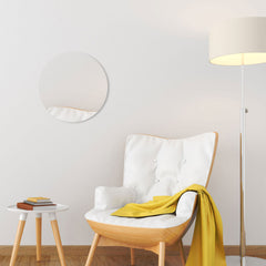 En modern läshörna med en vit stoppad stol med en gul filt, ett runt sidobord med böcker, en hög golvlampa och en Bozetto Spegel Rund Cirkel 50 cm på den vita väggen.