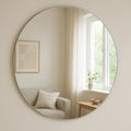 En Spegel Rund Cirkel 80 cm från Bozetto speglar ett mysigt vardagsrum med en ljus soffa, abstrakt väggkonst, sidobord i trä, krukväxt och skira gardiner som släpper in mjukt naturligt ljus.