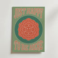 Just Happy Flower Poster by Bozetto föreställer en leende orange blomma som utstrålar enkel glädje, omgiven av grönt, med fet beige text ovanför och under: BARA GLAD ATT VARA HÄR.